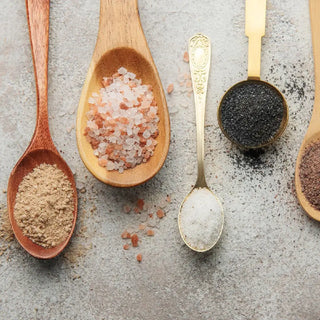 A variety of differ types of spoons laying on a table with herbs, spices and salts.