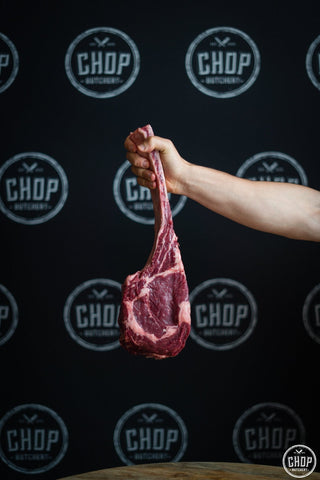 A hand holds up a large Tomahawk Steak (Minimum 1.6kg) against a black background with repeating CHOP Butchery logos—perfect for your next BBQ.